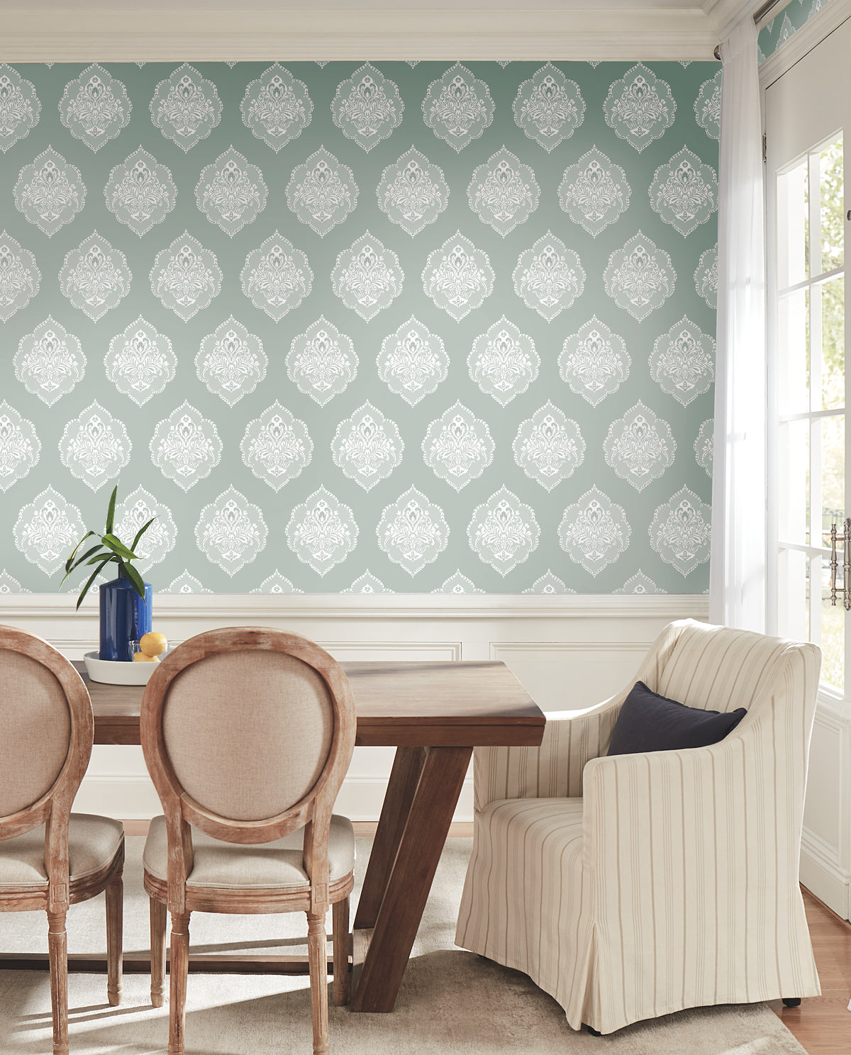 A dining room featuring a wooden table, three upholstered chairs, and a striped armchair is adorned with York Wallcoverings’ Signet Medallion Damask Wallpaper in blue. Sunlight pours through the window, illuminating a vase of green stems atop the table.