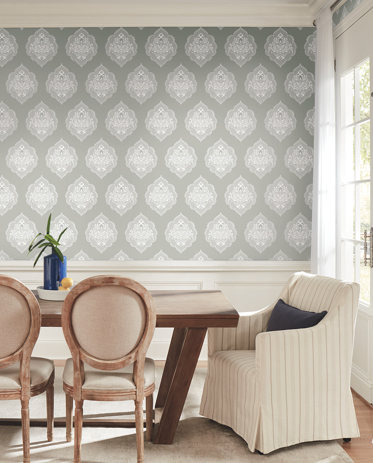 The dining room features a wooden table surrounded by three round-back chairs and a striped armchair, complemented by a vase filled with green leaves. Sunlight streams in from windows on the right, illuminating the walls adorned with York Wallcoverings’ Signet Medallion Damask Wallpaper in gray and white.