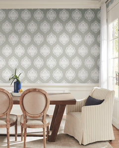 The dining room features a wooden table surrounded by three round-back chairs and a striped armchair, complemented by a vase filled with green leaves. Sunlight streams in from windows on the right, illuminating the walls adorned with York Wallcoverings’ Signet Medallion Damask Wallpaper in gray and white.