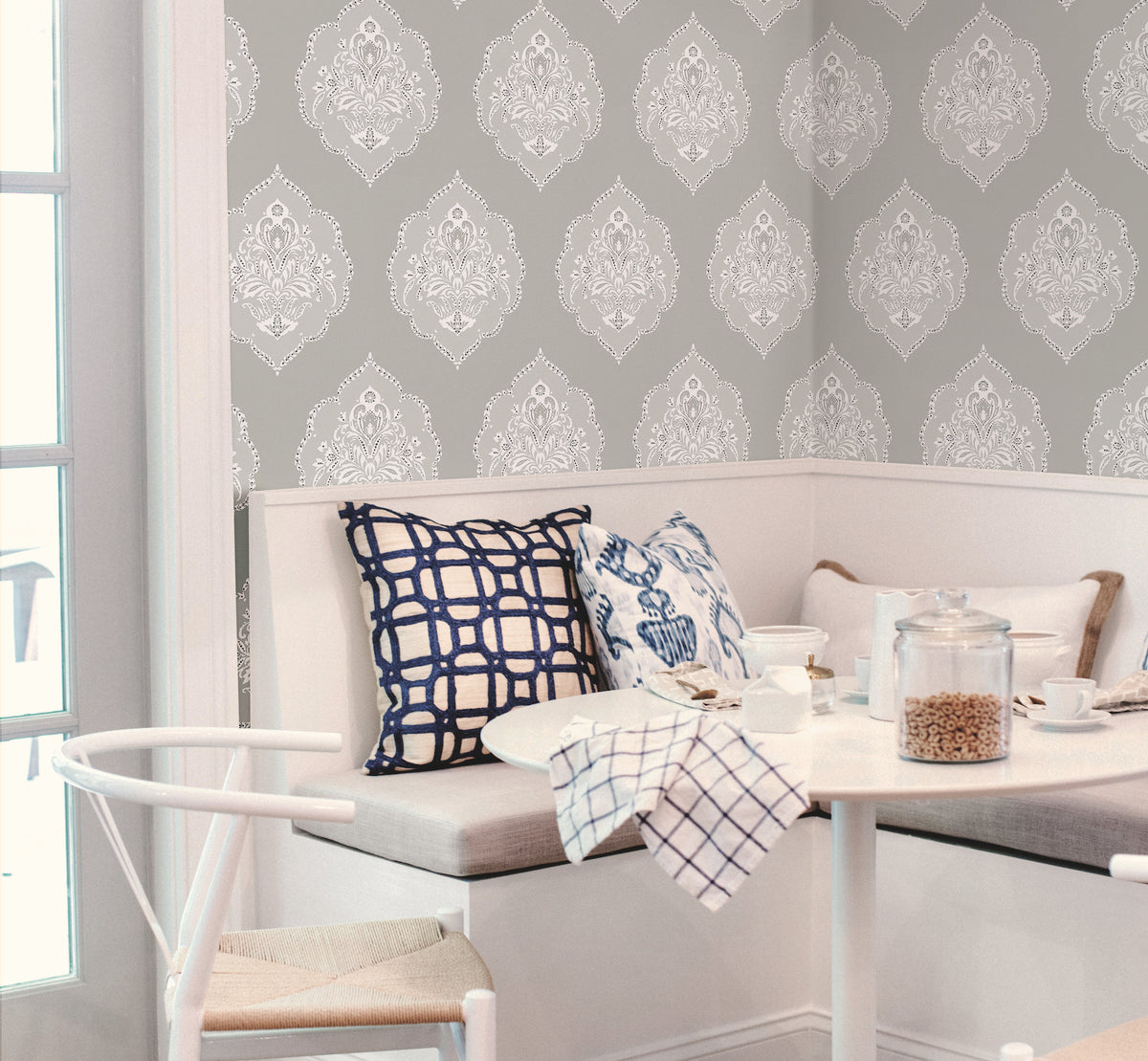 A cozy breakfast nook features a white bench, blue and white patterned pillows, a round table set for breakfast, and a backdrop of Signet Medallion Damask Wallpaper by York Wallcoverings in gray and white.