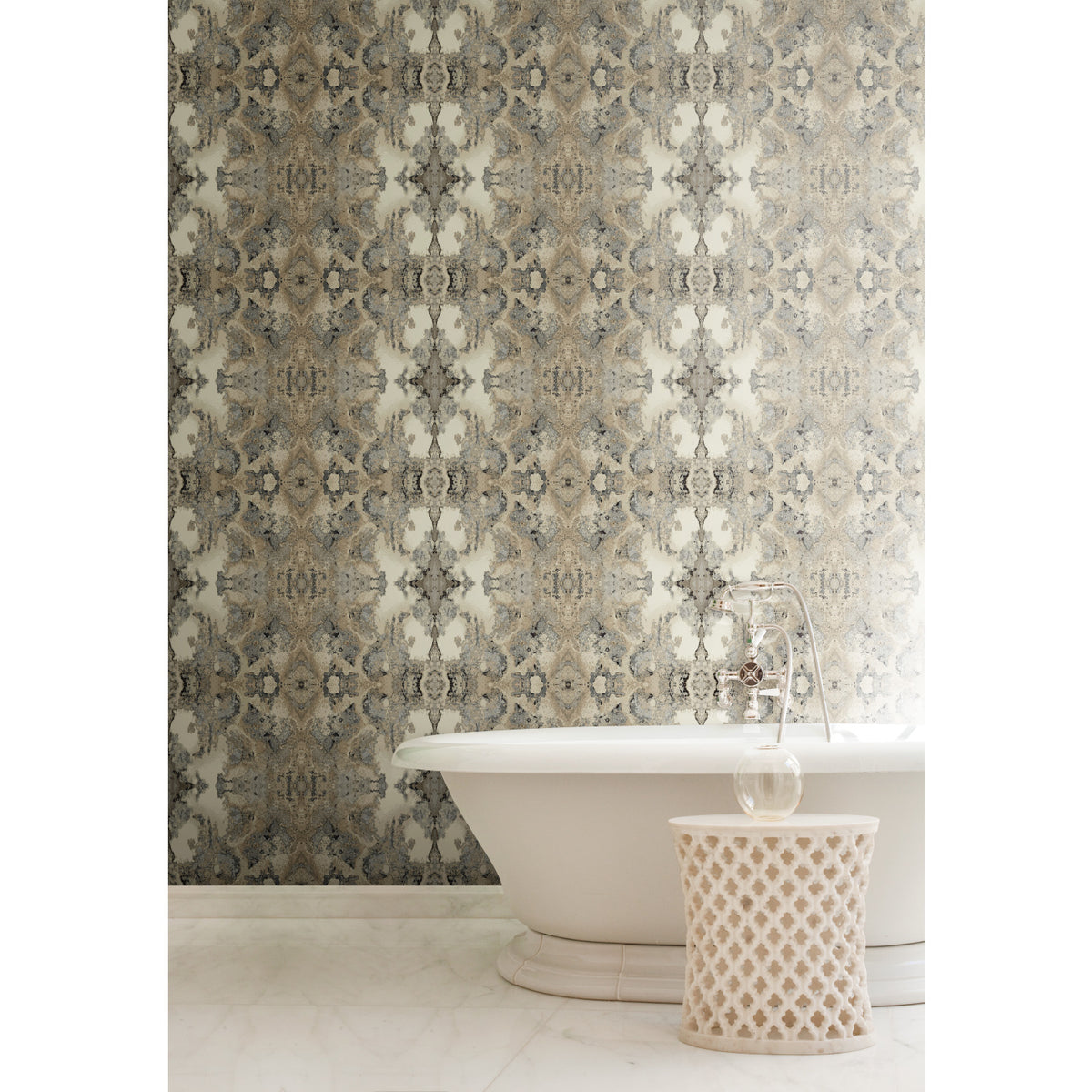 Inner Beauty Wallpaper by York Wallcoverings features an intricate, symmetrical damask-style pattern in shades of gray, beige, and gold, installed behind a white freestanding bathtub and a small, perforated side table.