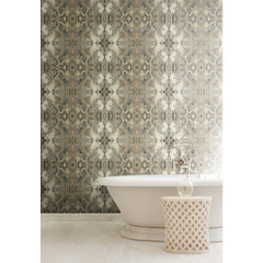 Inner Beauty Wallpaper by York Wallcoverings features an intricate, symmetrical damask-style pattern in shades of gray, beige, and gold, installed behind a white freestanding bathtub and a small, perforated side table.