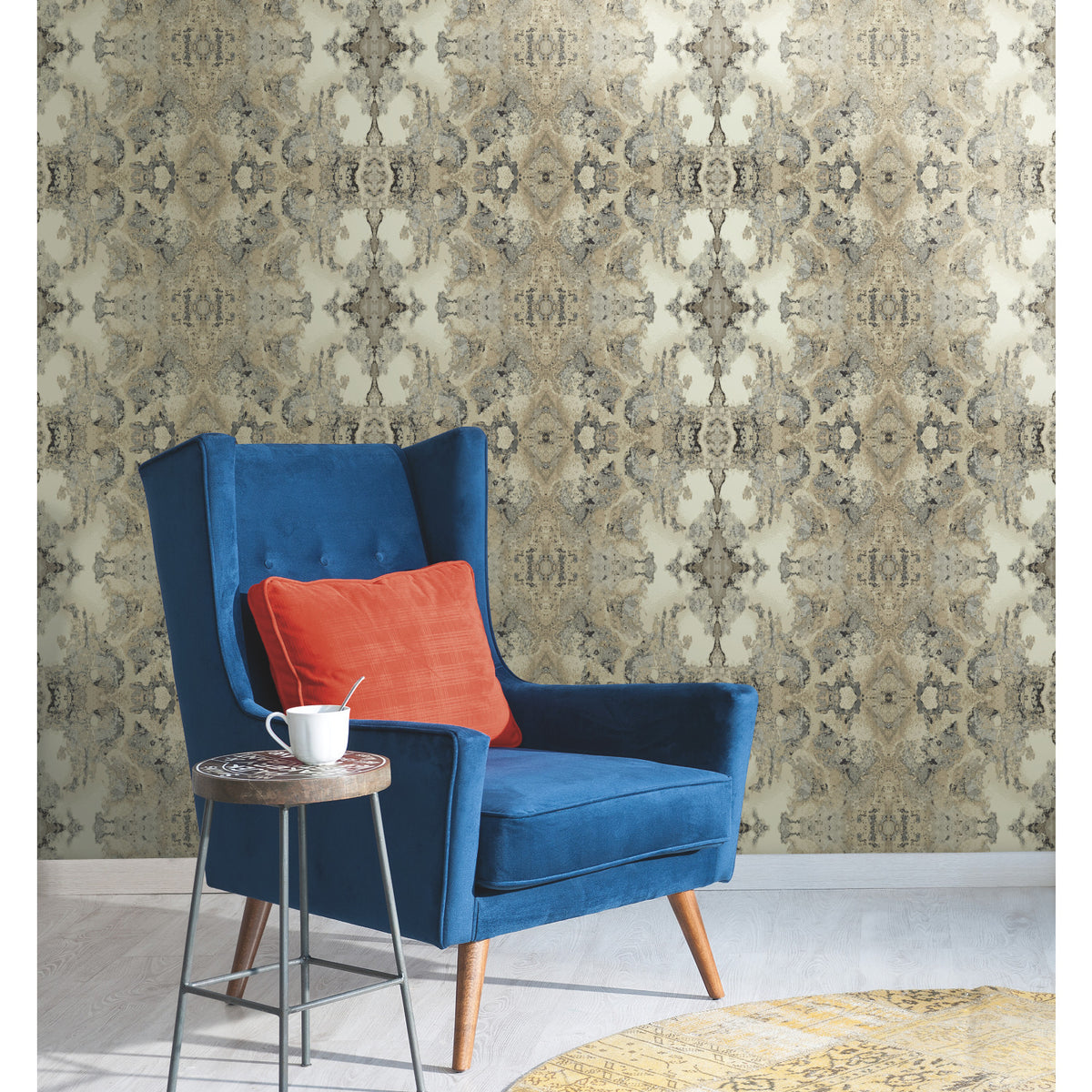 A blue wingback chair with an orange pillow sits in front of a wall covered in Inner Beauty Wallpaper by York Wallcoverings, featuring an ornate, repeating gray and cream pattern.