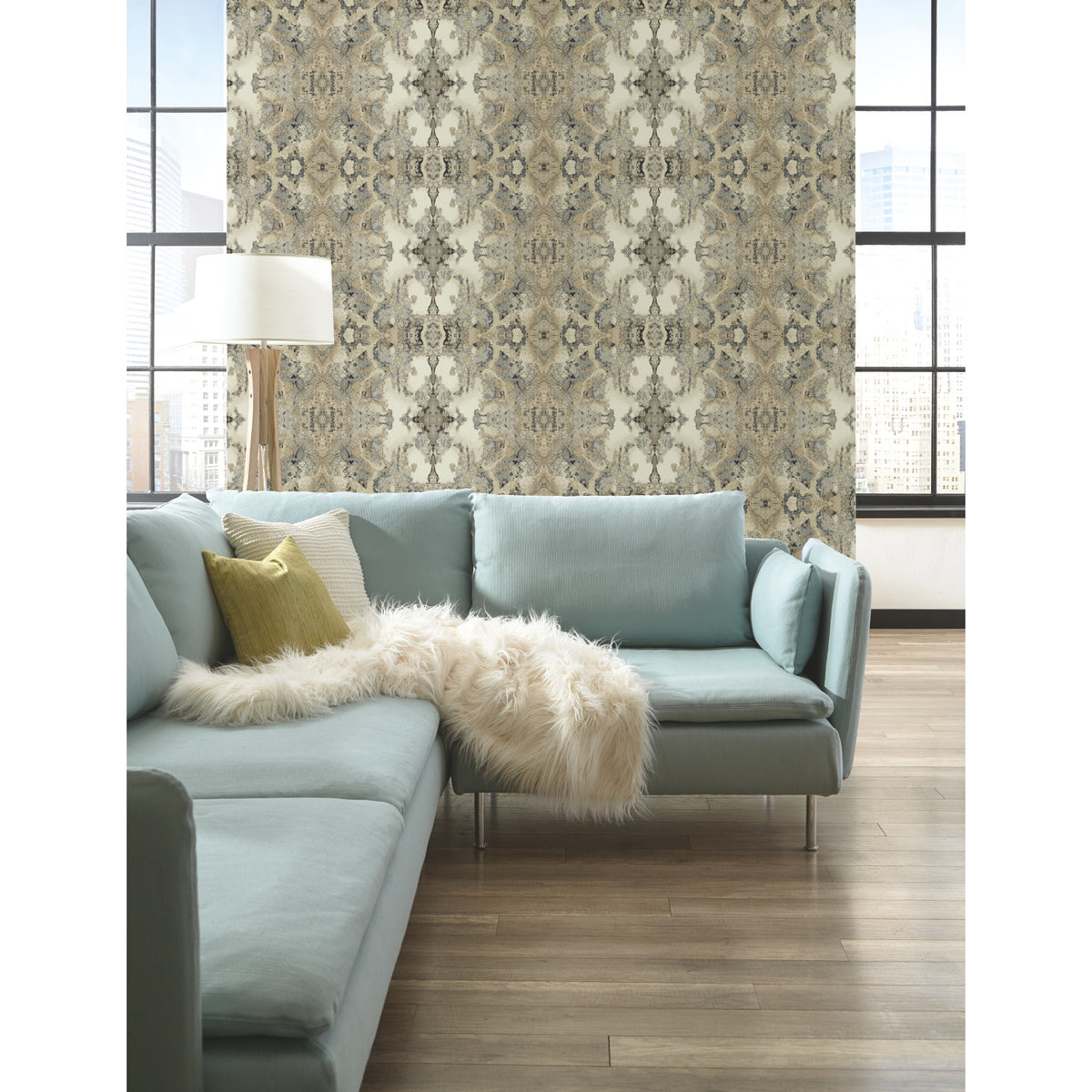 Inner Beauty Wallpaper by York Wallcoverings featuring a symmetrical, intricate brown and grey botanical pattern on a light background, displayed on a wall behind a light blue sectional sofa.