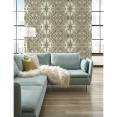 Inner Beauty Wallpaper by York Wallcoverings featuring a symmetrical, intricate brown and grey botanical pattern on a light background, displayed on a wall behind a light blue sectional sofa.