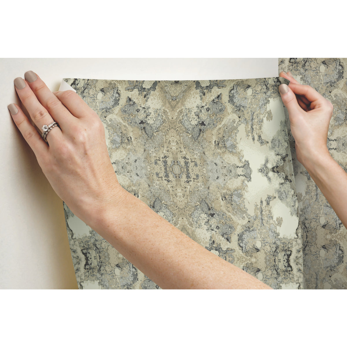 Two hands applying Inner Beauty Wallpaper, featuring a gray and beige stone-like marbled pattern, to a wall.