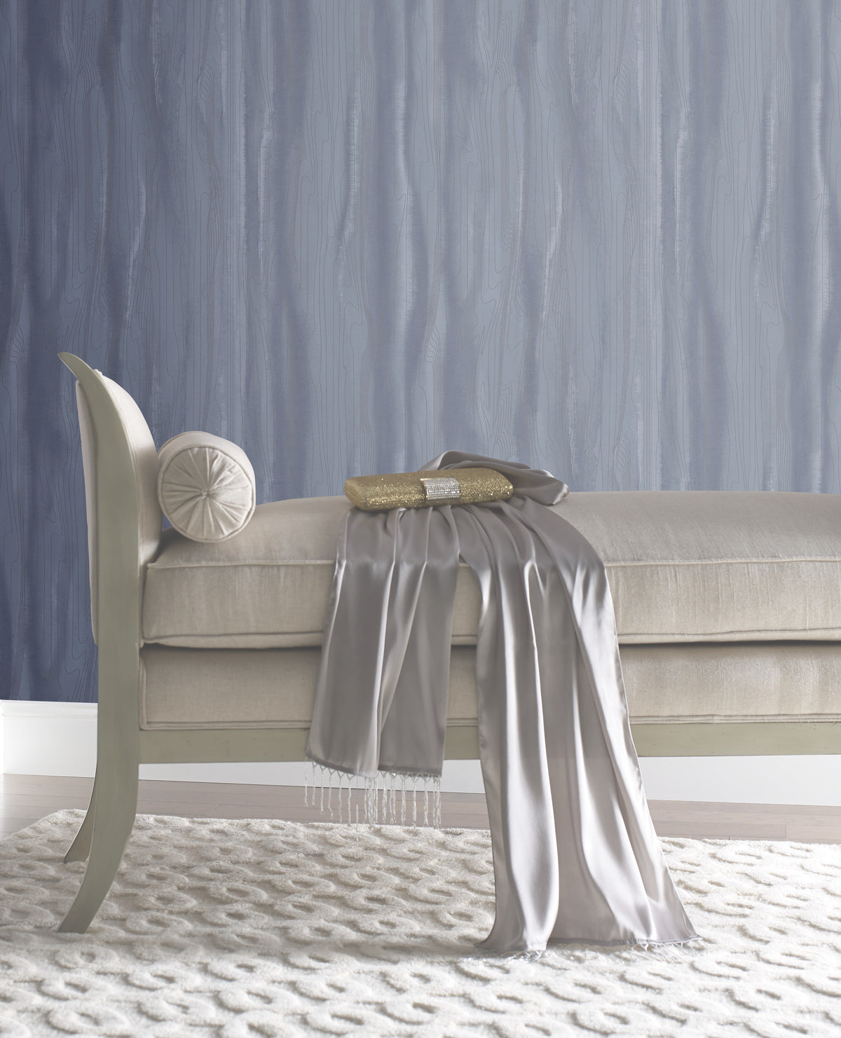 A beige upholstered bench with a round bolster cushion, a gold clutch, and a draped silver scarf, set against Candice Olson Fantasy Faux Bois Wallpaper by York Wallcoverings.