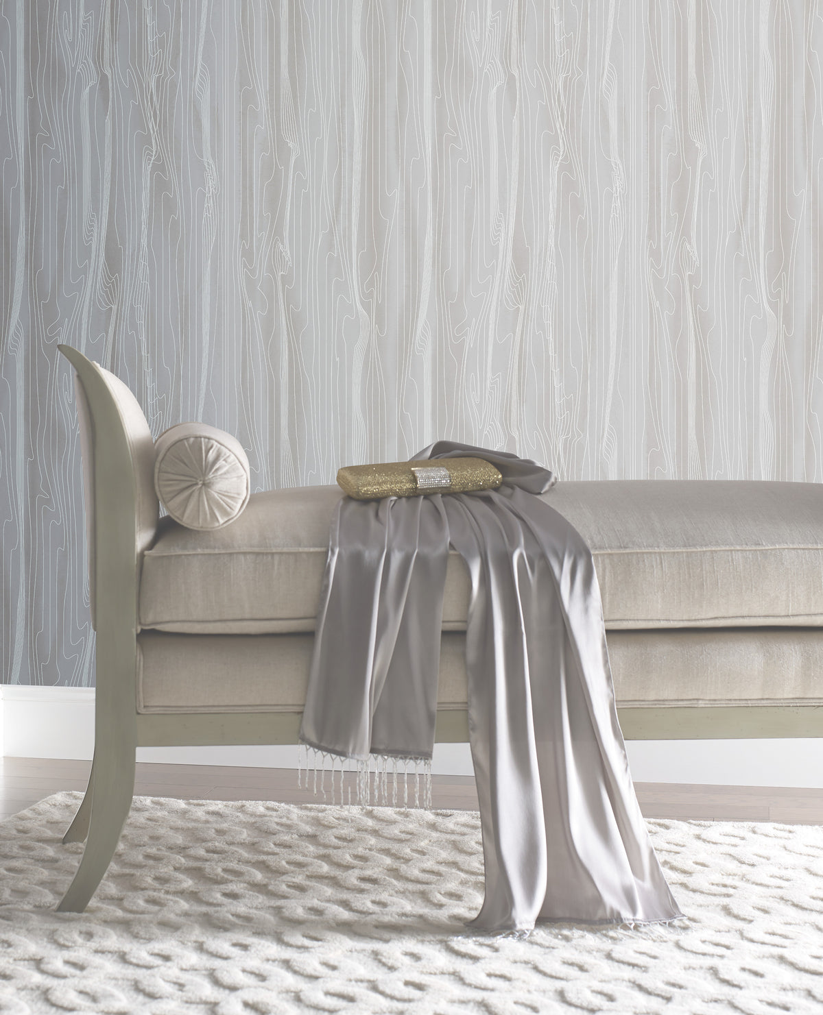 An upholstered bench with a small bolster pillow, a gold clutch, and a draped gray scarf, set against York Wallcoverings Candice Olson Fantasy Faux Bois Wallpaper featuring a white wood-grain pattern.