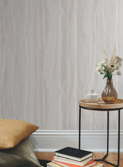Candice Olson Fantasy Faux Bois Wallpaper in light gray with a linear wood grain pattern displayed behind a side table with a glass of water and a floral arrangement.