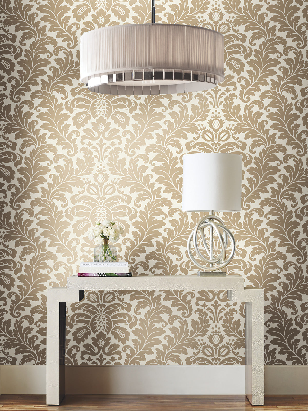 Candice Olson Modern Romance Wallpaper in an intricate cream and tan damask pattern installed behind a console table styled with a lamp, a small vase of flowers, and stacked books.