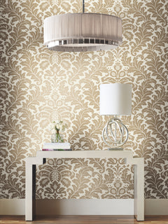 Candice Olson Modern Romance Wallpaper in an intricate cream and tan damask pattern installed behind a console table styled with a lamp, a small vase of flowers, and stacked books.