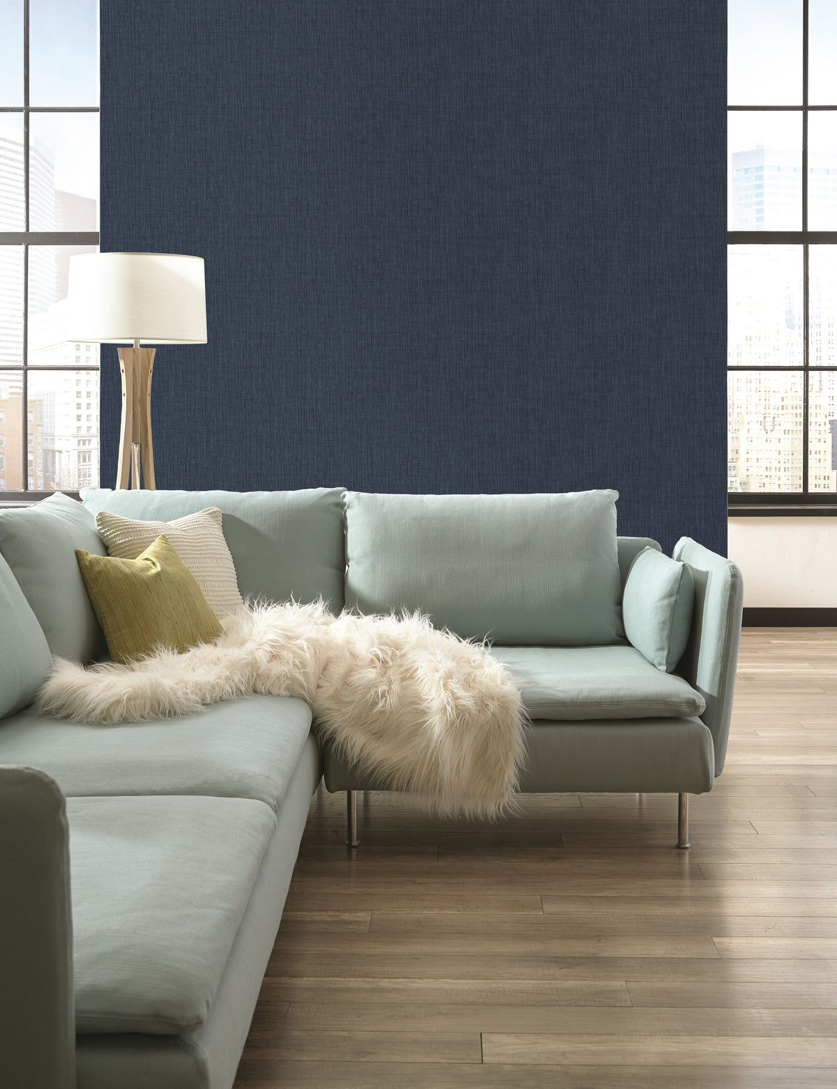 A room featuring dark navy Sweet Grass Wallpaper by York Wallcoverings, a light green sectional sofa, a floor lamp, and two large windows overlooking a city skyline.