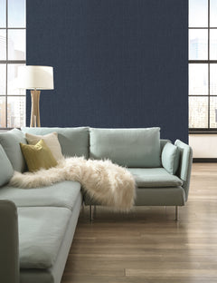 A room featuring dark navy Sweet Grass Wallpaper by York Wallcoverings, a light green sectional sofa, a floor lamp, and two large windows overlooking a city skyline.