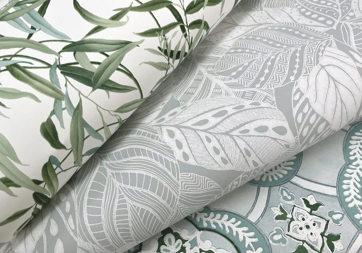 Three rolled sheets of Willow Grove Wallpaper by York Wallcoverings: one showcases green leafy branches, another displays intricate gray and white leaves, and the third features a blue and white floral pattern.