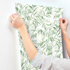 A person applies York Wallcoverings’ Willow Grove Wallpaper, featuring a green leafy vine pattern, to a white wall. Only their hands and arms—clad in a striped shirt with a ring—are visible.