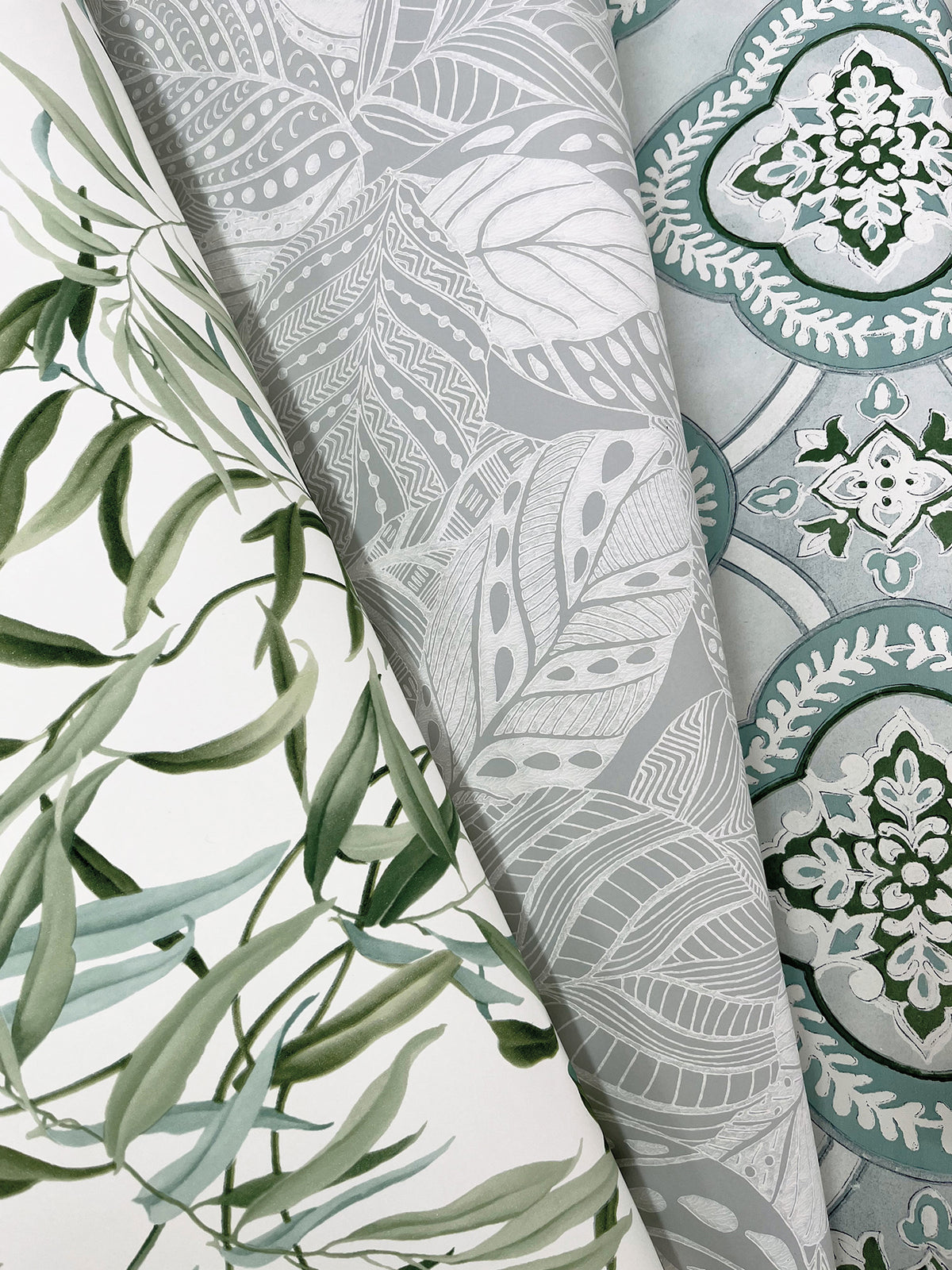 Three overlapping rolls of York Wallcoverings’ Willow Grove Wallpaper: one featuring green leaves, another with a gray and white abstract leafy pattern, and a third showcasing a blue and white ornate geometric design with floral motifs.