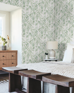 A bedroom features Willow Grove Wallpaper by York Wallcoverings, a wooden bench, and a dresser. White bedding, a textured blanket, a lamp, and sunlit windows complete the cozy, modern decor.