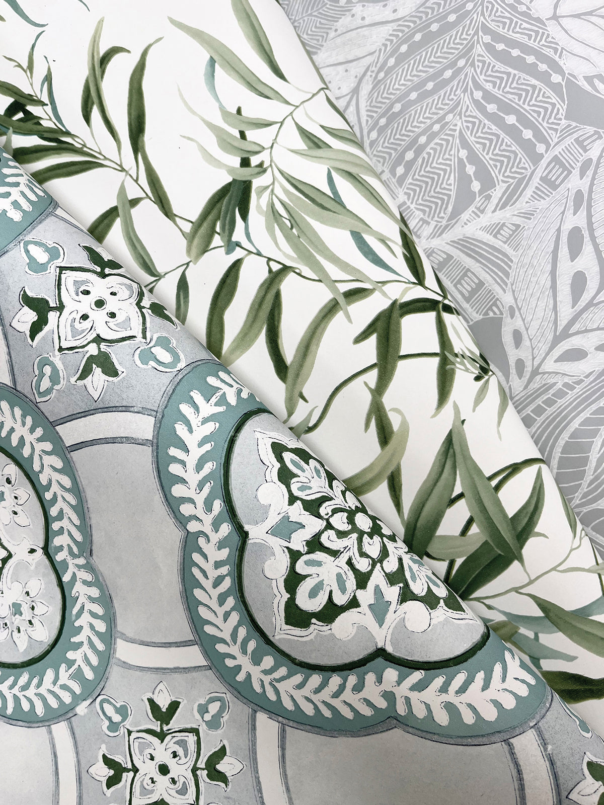 Three different patterned wallpapers overlap: York Wallcoverings’ Willow Grove Wallpaper featuring green leafy branches, one with light gray intricate abstract designs, and another with teal and white ornate floral motifs set against a gray background.