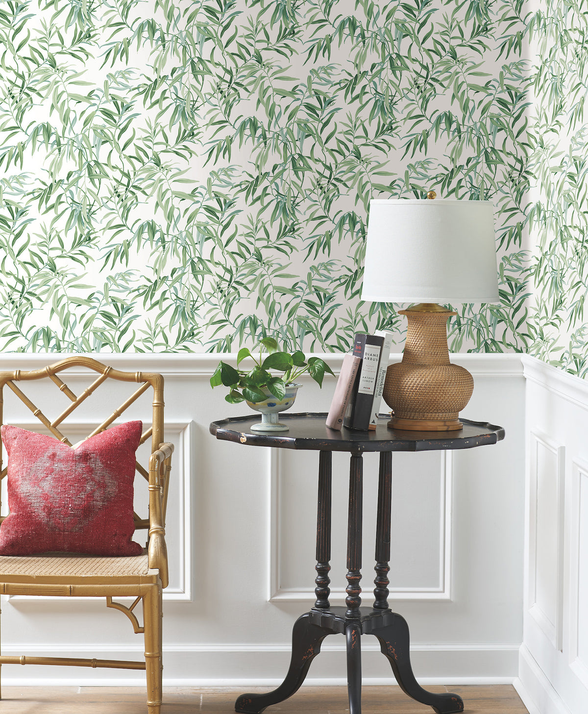 A small round table with books, a potted plant, and a lamp sits beside a wooden chair with a red pillow. The background features Willow Grove Wallpaper by York Wallcoverings and white wall paneling.