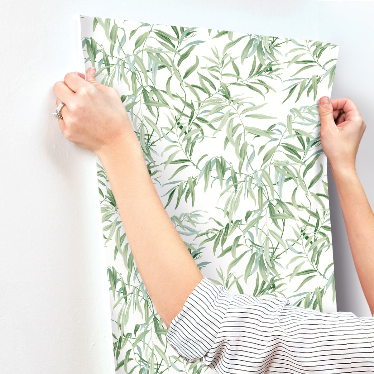 A person wearing a striped shirt hangs Willow Grove Wallpaper by York Wallcoverings, featuring a leafy green pattern, on a white wall.