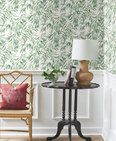 A wooden chair with a red pillow sits beside a round black table holding a plant, books, and a wicker lamp. The background features Willow Grove Wallpaper by York Wallcoverings with green leafy patterns above classic white paneling.