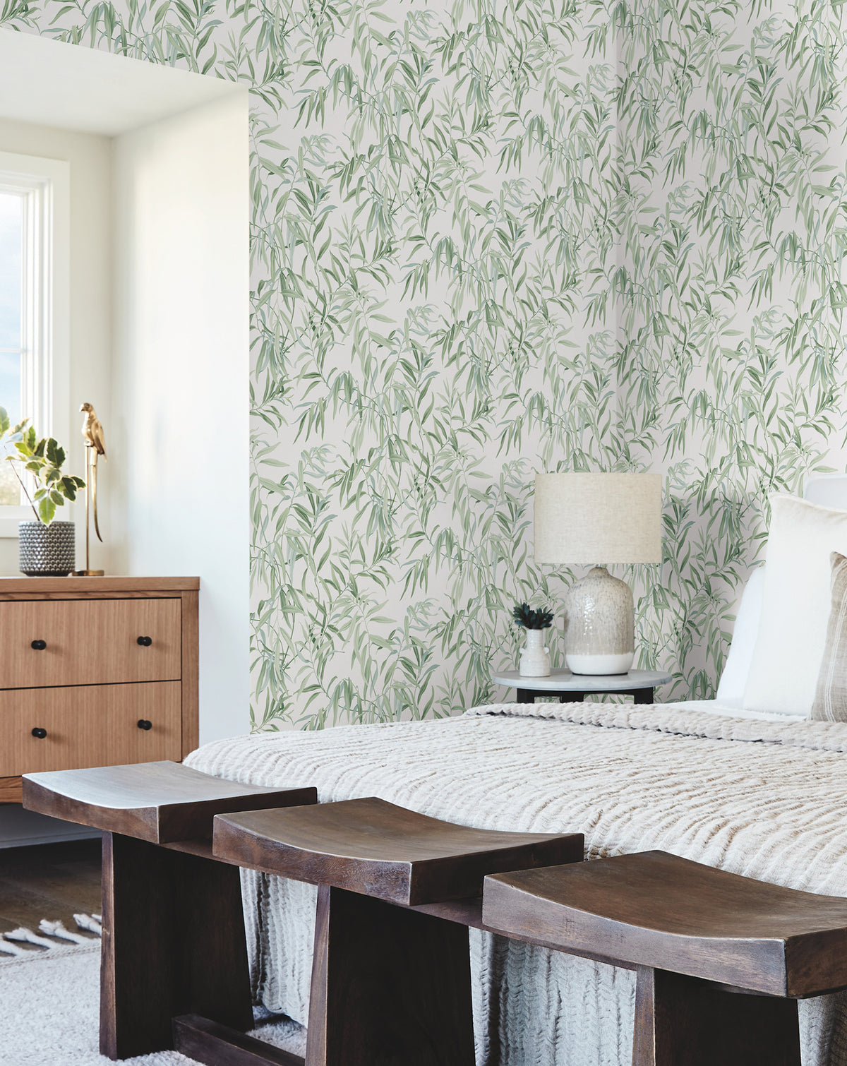 Bedroom featuring Willow Grove Wallpaper by York Wallcoverings, a wooden bed adorned with pillows and a lamp, and dark benches. A dresser with decor sits beside a window that fills the space with natural light, creating a cozy and modern atmosphere.