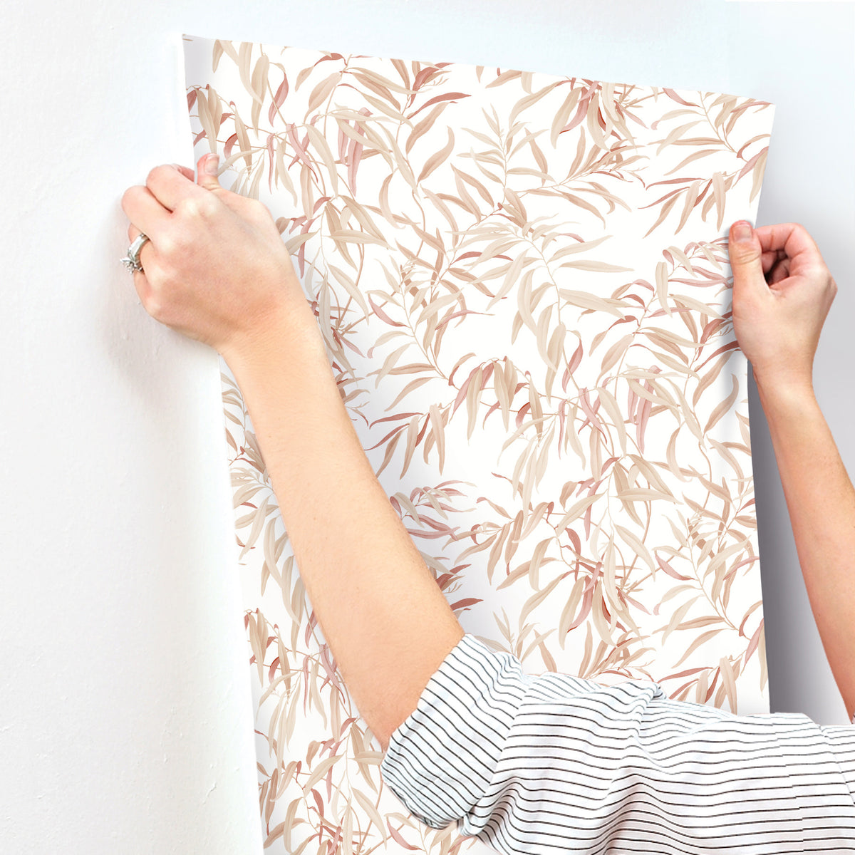 A person in a striped shirt applies York Wallcoverings’ Willow Grove Wallpaper, featuring a beige and light brown leafy pattern, to a white wall.