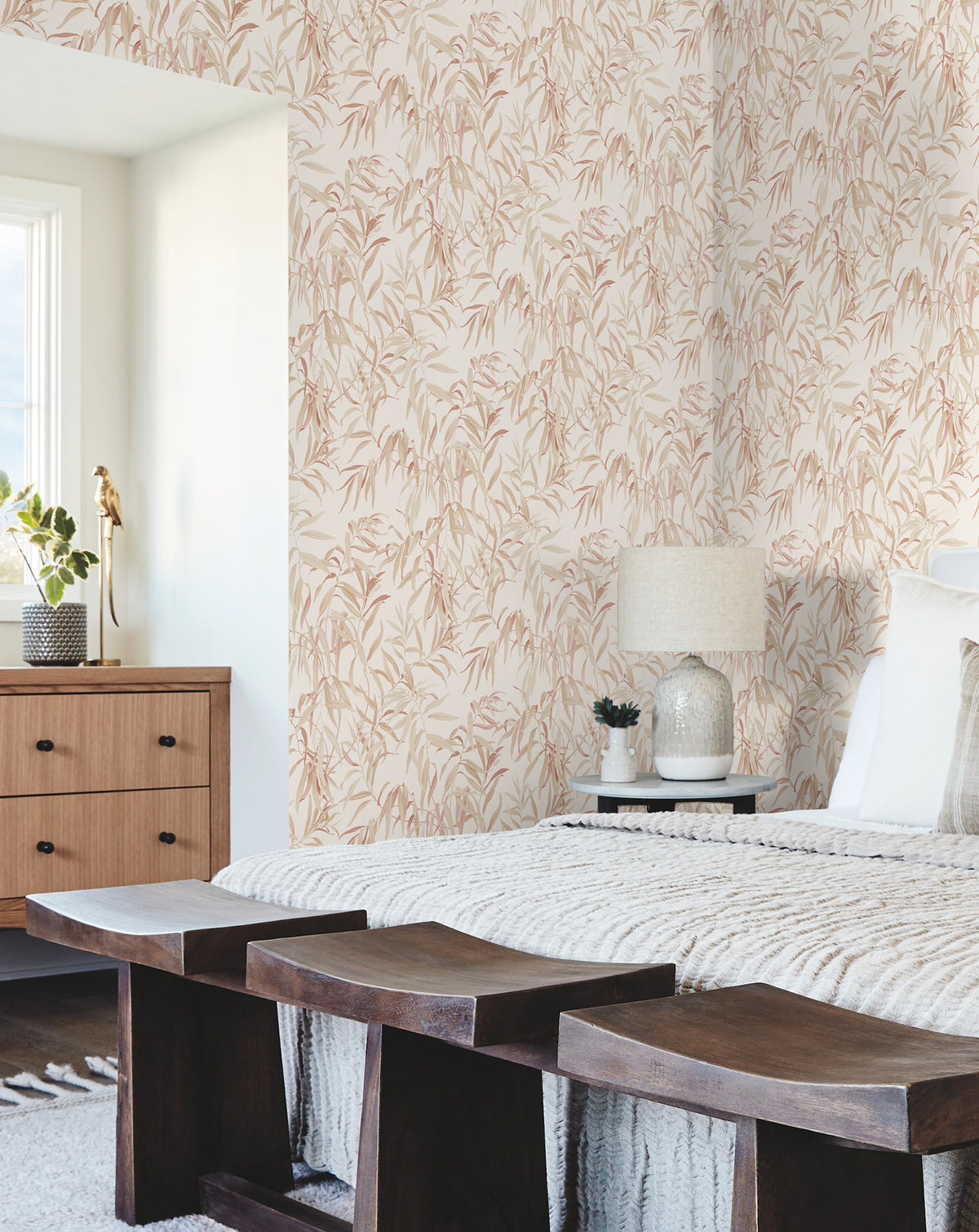 A cozy bedroom featuring Willow Grove Wallpaper by York Wallcoverings, a neatly made bed, wooden benches at the foot, a wooden dresser with a vase, and a lamp resting on the nightstand. Sunlight streams in through the window on the left.