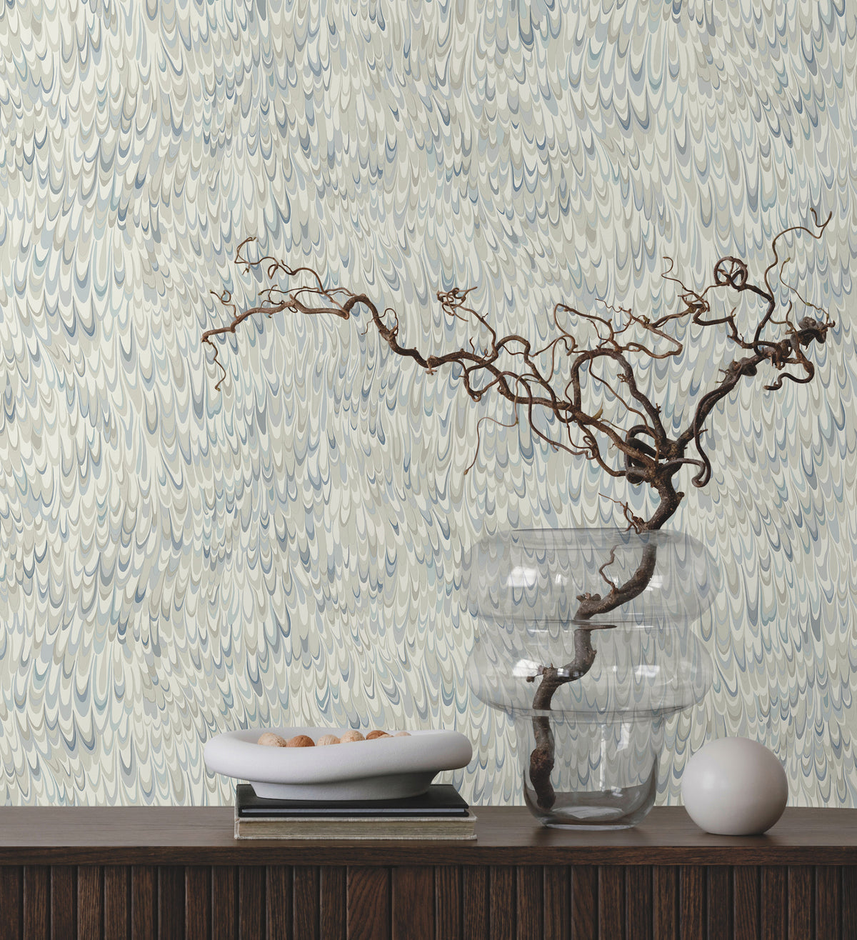 Ronald Redding Aslan Wallpaper featuring an intricate pattern of overlapping blue and grey petal-like shapes, displayed behind a shelf with a clear vase, a bowl of small stones, and a decorative orb.
