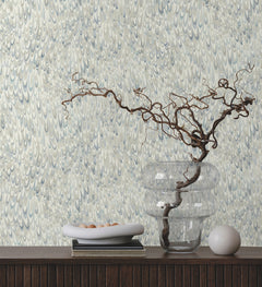 Ronald Redding Aslan Wallpaper featuring an intricate pattern of overlapping blue and grey petal-like shapes, displayed behind a shelf with a clear vase, a bowl of small stones, and a decorative orb.