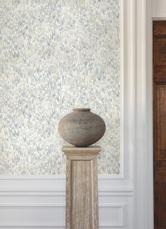 Ronald Redding Aslan Wallpaper featuring a repeating pattern of pale blue and tan teardrop shapes on a white background, displayed behind a ceramic vessel on a wooden pedestal.