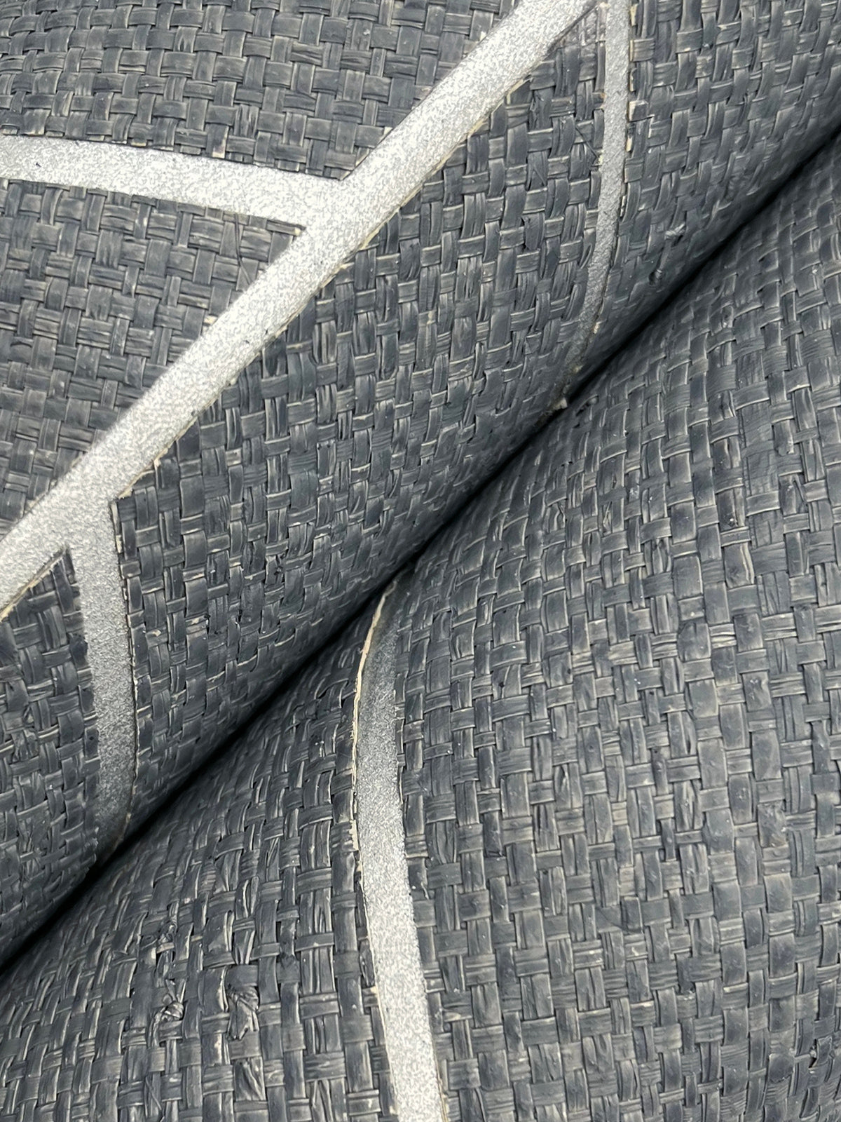 Close-up of two rolls of Ronald Redding Atelier Herringbone Wallpaper by York Wallcoverings, featuring a textured, dark gray surface with a woven pattern and thin, light gray diagonal lines.
