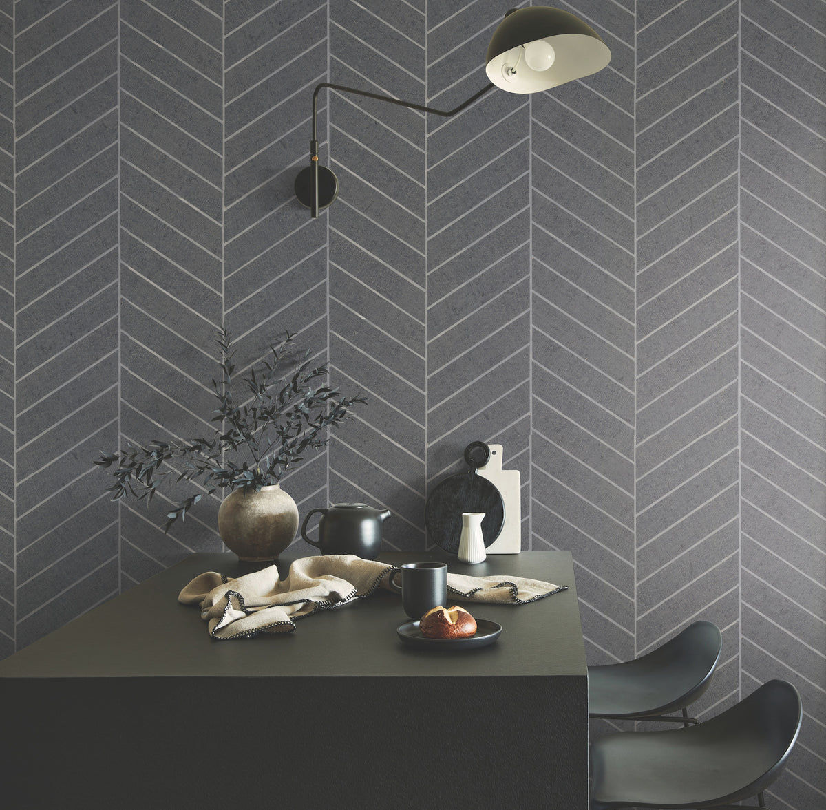 Modern dining area featuring a black table, two black chairs, breakfast items, and a vase with branches set against a grey herringbone wall adorned with Ronald Redding Atelier Herringbone Wallpaper by York Wallcoverings. Overhead, a black wall lamp provides light.