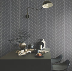 Modern dining area featuring a black table, two black chairs, breakfast items, and a vase with branches set against a grey herringbone wall adorned with Ronald Redding Atelier Herringbone Wallpaper by York Wallcoverings. Overhead, a black wall lamp provides light.