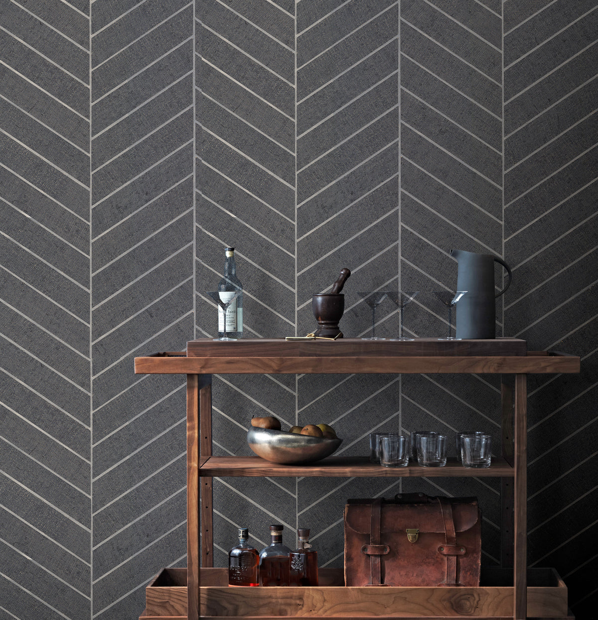 A wooden bar cart with liquor bottles, a metal bowl filled with fruit, glasses, a leather bag, and a pitcher sits against a wall adorned with Ronald Redding Atelier Herringbone Wallpaper by York Wallcoverings.