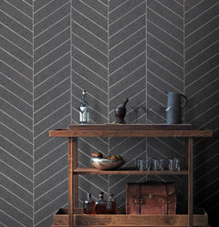 A wooden bar cart with liquor bottles, a metal bowl filled with fruit, glasses, a leather bag, and a pitcher sits against a wall adorned with Ronald Redding Atelier Herringbone Wallpaper by York Wallcoverings.