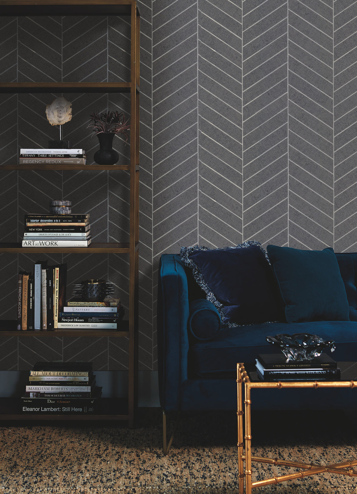 A modern living room featuring a dark blue velvet sofa, a gold-accented coffee table, a bookshelf filled with books and decor, and gray Ronald Redding Atelier Herringbone Wallpaper by York Wallcoverings.