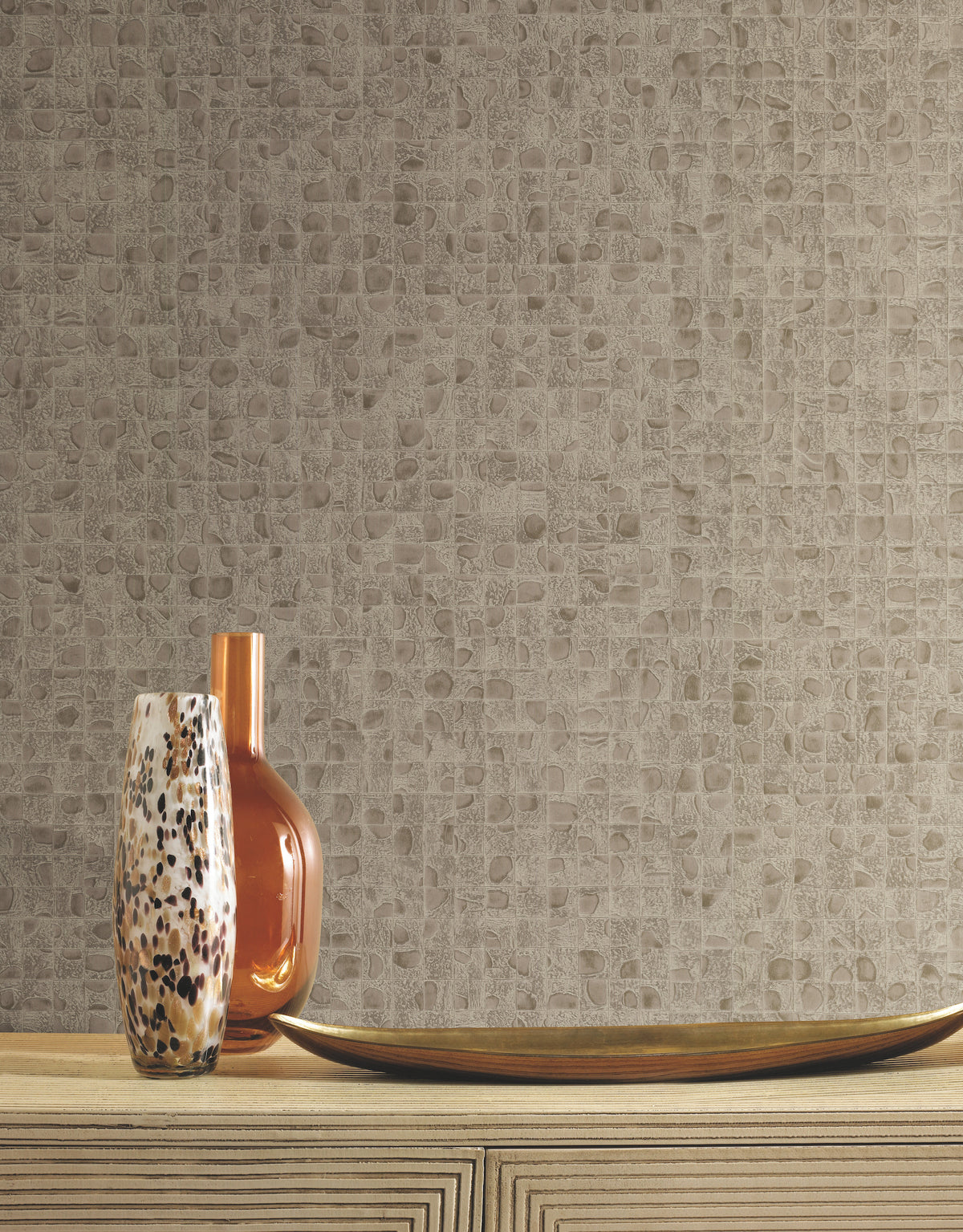 Two glass vases and a long, curved decorative dish rest on a wooden console table against a neutral-toned Ronald Redding Leather Lux Wallpaper with a textured, mosaic-like grid pattern.