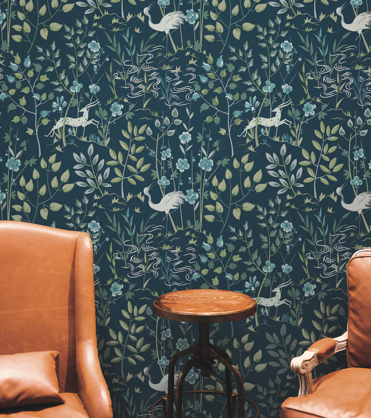 Ronald Redding Aspen Wallpaper by York Wallcoverings features a repeating pattern of white deer, cranes, and floral sprigs on a dark blue background, positioned behind two brown leather chairs and a small wooden side table.