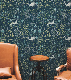 Ronald Redding Aspen Wallpaper by York Wallcoverings features a repeating pattern of white deer, cranes, and floral sprigs on a dark blue background, positioned behind two brown leather chairs and a small wooden side table.
