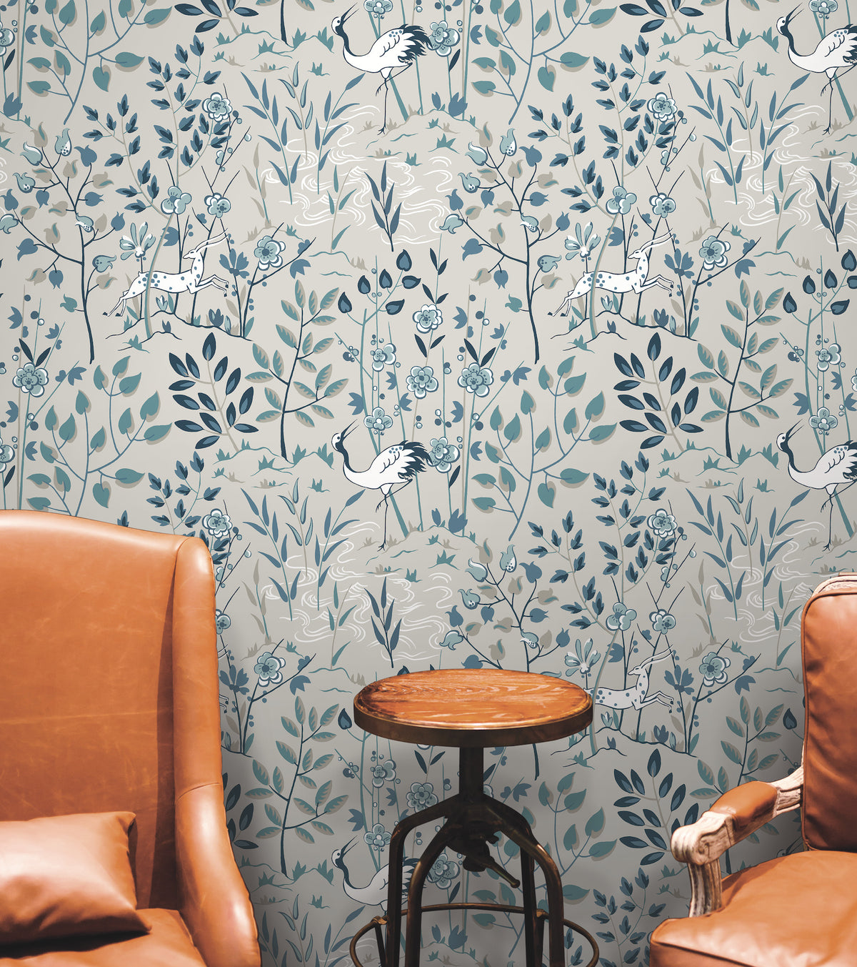 Ronald Redding Aspen Wallpaper featuring stylized cranes and spotted deer among blue and grey foliage on a light background, positioned between two leather chairs and a small round table.