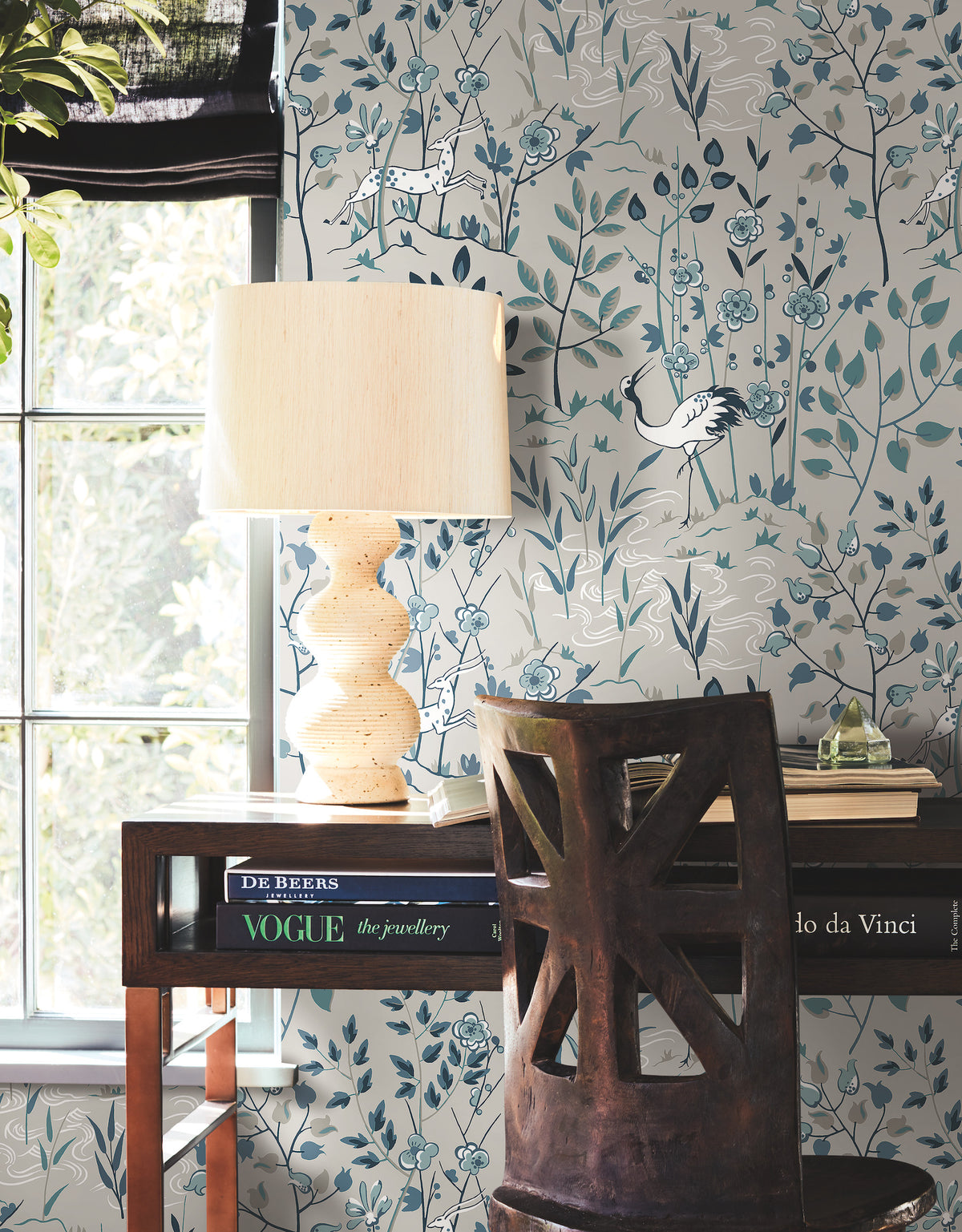Ronald Redding Aspen Wallpaper featuring an illustrated scene of cranes, stylized trees, and flowers in shades of blue, white, and sage green, installed in a home office with a desk and lamp.