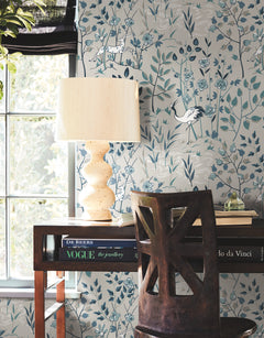 Ronald Redding Aspen Wallpaper featuring an illustrated scene of cranes, stylized trees, and flowers in shades of blue, white, and sage green, installed in a home office with a desk and lamp.