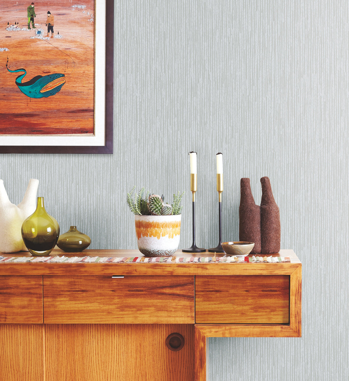 Ronald Redding Feather Fletch Wallpaper in light grey with a vertical, feathered texture displayed behind a wooden sideboard holding vases, a potted cactus, and candle holders.