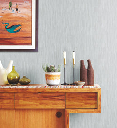 Ronald Redding Feather Fletch Wallpaper in light grey with a vertical, feathered texture displayed behind a wooden sideboard holding vases, a potted cactus, and candle holders.