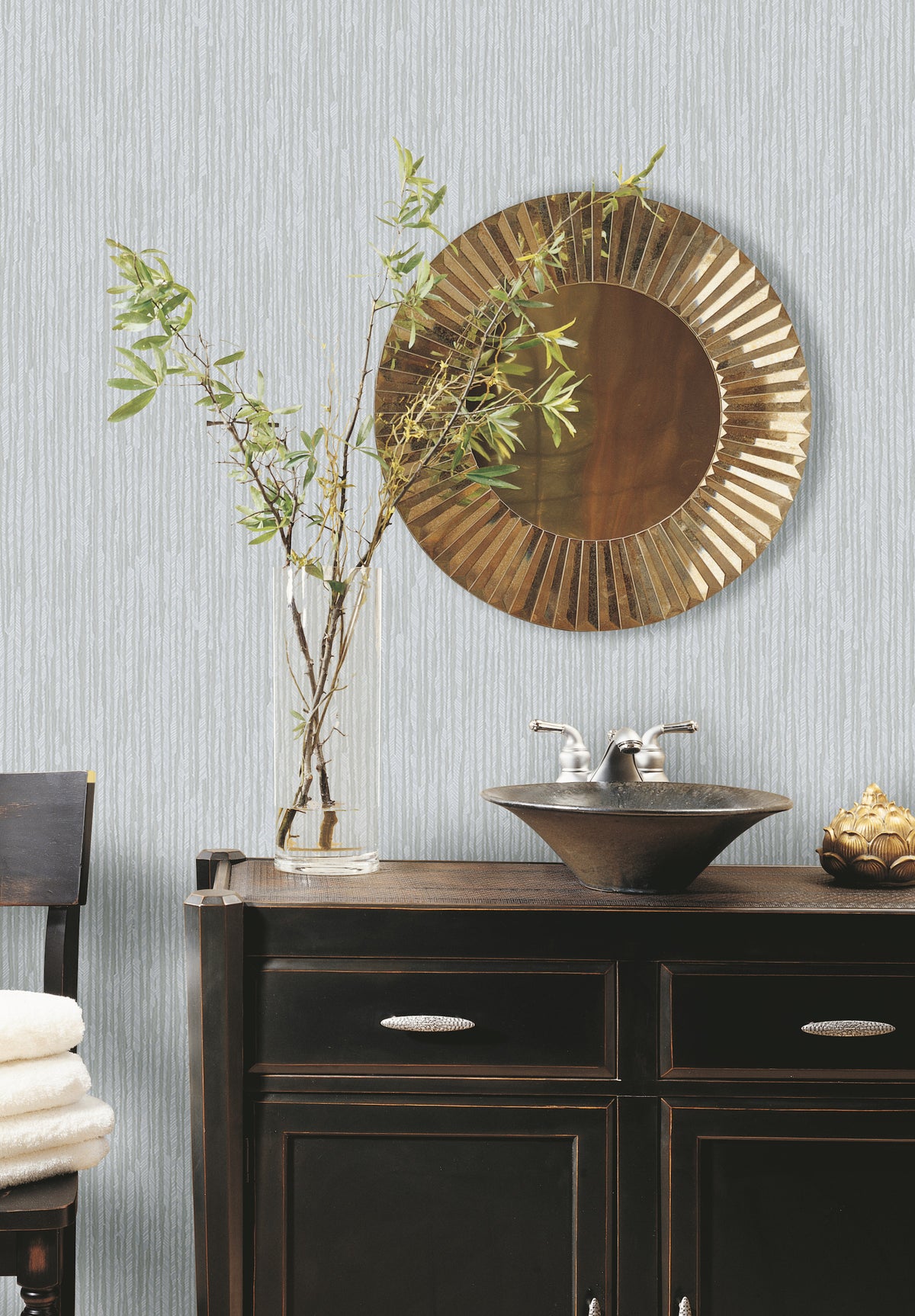 A bathroom vanity with a vessel sink and a round, textured metallic mirror set against light blue, vertical-striped Ronald Redding Feather Fletch Wallpaper by York Wallcoverings.