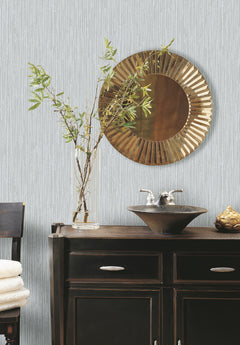 A bathroom vanity with a vessel sink and a round, textured metallic mirror set against light blue, vertical-striped Ronald Redding Feather Fletch Wallpaper by York Wallcoverings.