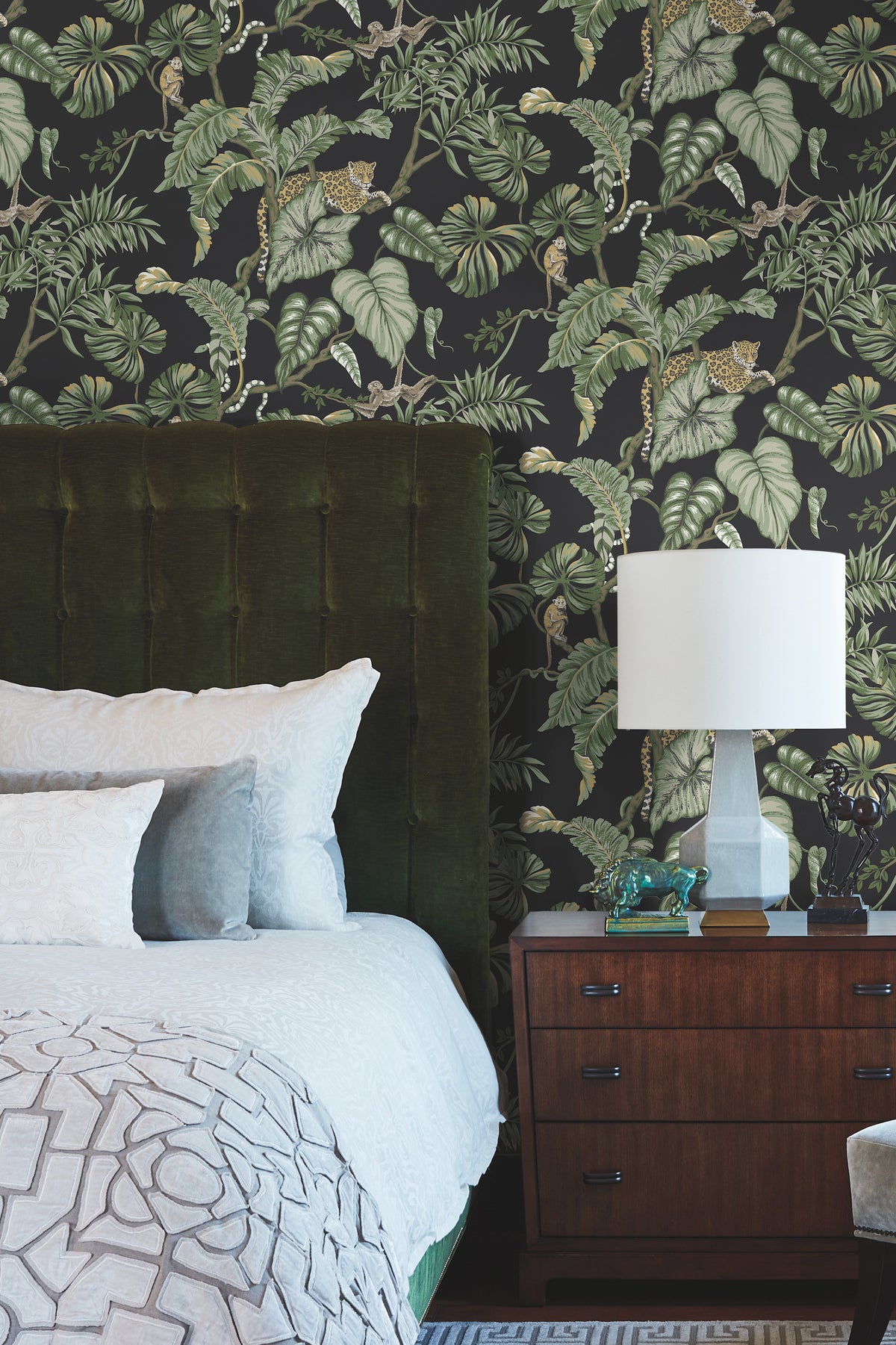 Bedroom featuring Ronald Redding Jungle Cat Wallpaper with a dark tropical jungle pattern of green foliage, monkeys, and leopards behind a bed with a dark green headboard and a wooden nightstand with a lamp.