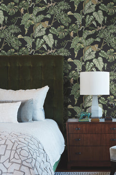 Bedroom featuring Ronald Redding Jungle Cat Wallpaper with a dark tropical jungle pattern of green foliage, monkeys, and leopards behind a bed with a dark green headboard and a wooden nightstand with a lamp.