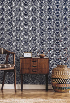 Ronald Redding Lotus Palm Wallpaper featuring a repeating dark blue and white stylized floral pattern behind a wooden nightstand, a chair, and a woven basket.